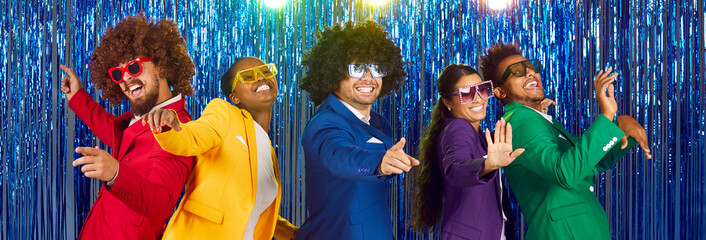 Panoramic photo of funny funny group of happy young people dressed in bright costumes, voluminous curly wigs and colorful big glasses, dancing and fooling around on sparkling blue background.