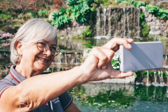 White-Haired Smiling Senior Woman Takes a Selfie with a Beautiful Waterfall in the Garden Enjoying Nature and Relax