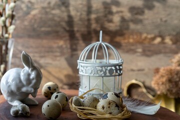 Quail eggs in a nest lying on a dark wooden surface, against the background of an Easter bunny made of white porcelain and a cage with feathers