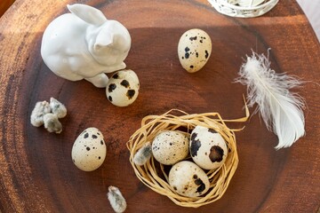 Quail eggs in a nest lying on a dark wooden surface, against the background of an Easter bunny made of white porcelain  