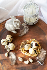 Quail eggs in a nest lying on a dark wooden surface, against the background of an Easter bunny made of white porcelain and a cage with feathers