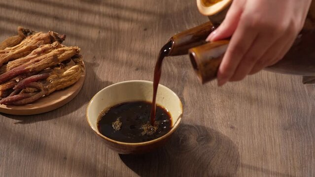 Female hand is pouring medicine from the bottle into a small bowl on wooden table background. Simulation scene for traditional Chinese medicine, decoction of red ginseng root.