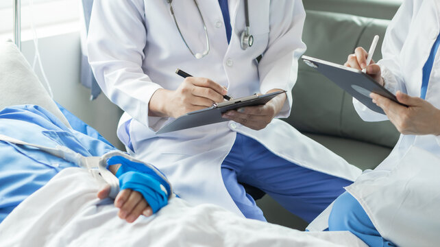 Cropped image of doctor in uniform with stethoscope using tablet standing in hospital with patient lying in bed on background Operator checks patient information on digital tablet