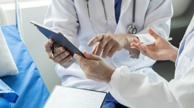 Cropped image of doctor in uniform with stethoscope using tablet standing in hospital with patient lying in bed on background Operator checks patient information on digital tablet