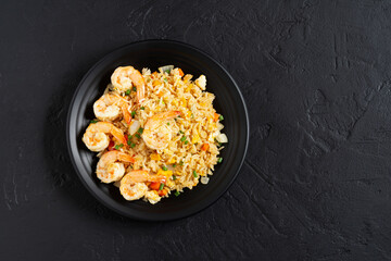 Fried rice with vegetables and sea prawns, carrots, eggs, and tomatoes. Thai Chinese food is arranged on a black plate on a black background kitchen table, top view.