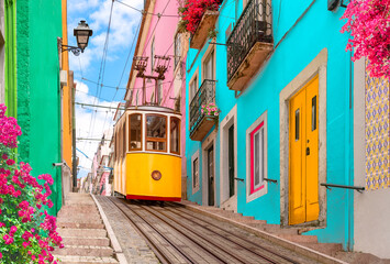 Lisbon, Portugal - Yellow tram on a street with colorful houses and flowers on the balconies - Bica Elevator going down the hill of Chiado. - obrazy, fototapety, plakaty