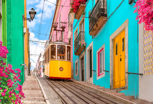 Lisbon, Portugal - Yellow tram on a street with colorful houses and flowers on the balconies - Bica Elevator going down the hill of Chiado.