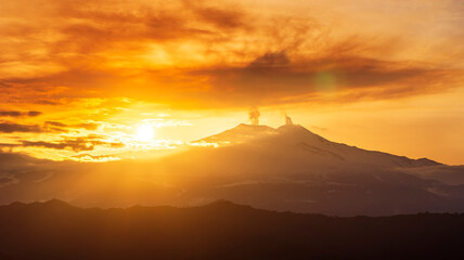 mysterious landscape of great erupting volcano with smoke from craters and snow on slopes in orange...