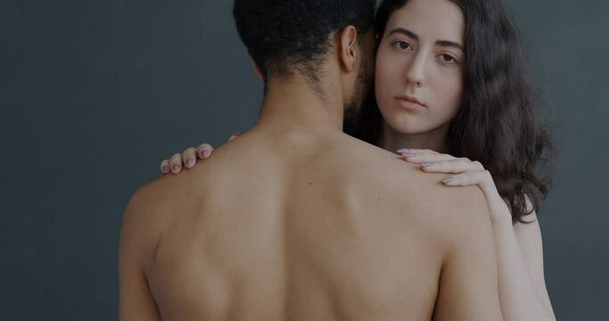 Slow motion of young woman hugging topless man and looking at camera with serious face on grey background. Love and affection concept.