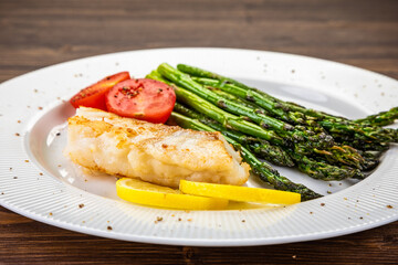 Fish dish - fried cod with green asparagus, lemon and fresh tomatoes on wooden table
