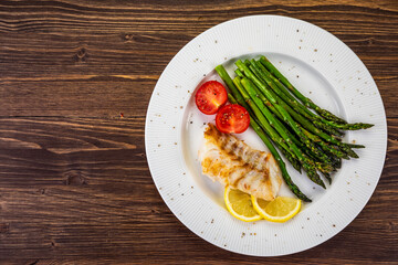 Fish dish - fried cod with green asparagus, lemon and fresh tomatoes on wooden table
