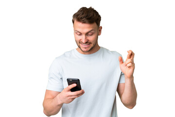 Young handsome caucasian man over isolated chroma key background using mobile phone with fingers crossing