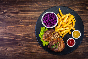 Fried beef sirloin with potatoes and vegetables on black stone plate on wooden table
