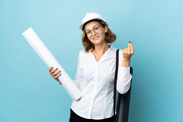 Young architect Georgian woman with helmet and holding blueprints over isolated background making money gesture