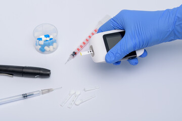 Trzymany w dłoniach w niebieskich rękawiczkach glukometr, obok strzykawki do insuliny, lekarstw - obrazy, fototapety, plakaty