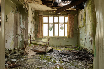 Derelict Hospital ward with collapsed ceiling and hole in roof, peeling paint and mould on the walls with upended broken table  and curtains still hanging.