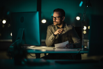 Portrait of tired young man working late at night at office with computer