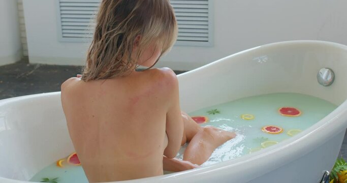 Sexy woman sitting in bathroom. blonde girl bathing in milk bath filled with fruit, relax spa skin care concept, back rare view slow motion, female touching her neck, shoulder sensuality sexualtity