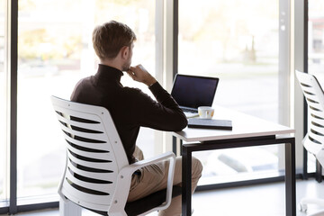 Pensive man sitting in office at table, working on laptop, back view, modern workplace
