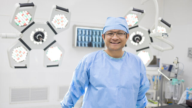 Asian surgeon or doctor smiling and standing inside operating room in hospital with blue uniform.Surgical lamp technology and MRI image on background.Kindness and happiness waiting for surgery.