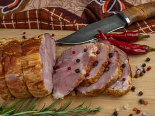 Sliced slices of juicy pork ham on a wooden cutting board, next to it are a carving knife, multi-colored allspice peas, red hot peppers, parsley, dill and basil. The concept of delicious meat products