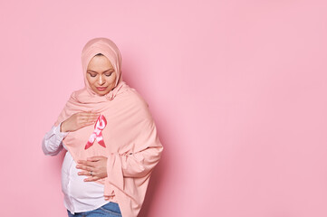 Muslim pregnant woman, holds her hands around pink satin awareness ribbon on hijab, shows her...