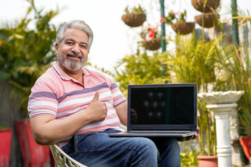 Indian senior man showing laptop screen while siting at garden.