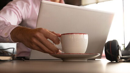 man's hand picking up a cup of coffee behind the laptop