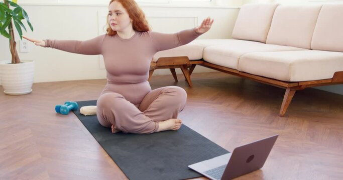 woman with long red hair doing physical exercise watching workout video on laptop, attractive girl following online training lesson, sitting on mat with crossed legs, performing under chin exercise