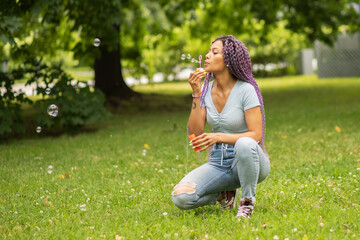 Naklejka na ściany i meble african american woman outdoor,a girl with colored hair in the summer in the park blows soap bubbles in nature,happiness and joy