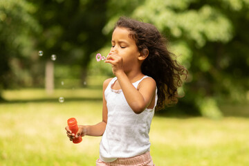 happy african american child in park, a girl plays in nature in the summer, a kid blows soap bubbles