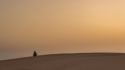 Fototapeta na wymiar una persona contemplando el amanecer sentada sobre la arena en una duna en el desierto