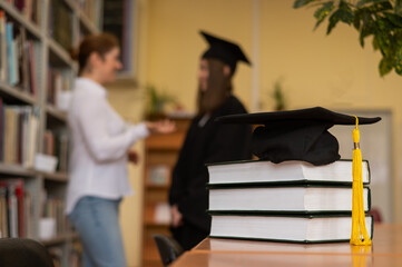 Close-up of a graduation cap on a stack of books in a library. teacher and young female graduate in...