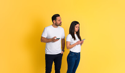 Possessive young adult man peeking at girlfriend's smart phone and trying to read her text messages while standing isolated on yellow background