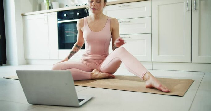 A smiling sporty girl in a pink workout tight suit is doing stretching in front laptop