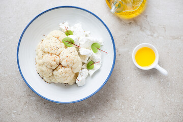 White plate with steamed cauliflower cabbage and feta cheese, flat lay on a beige stone background,...