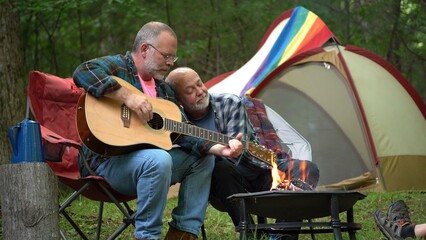 Two gay men enjoying nature camping with tent with pride flag in forest on river playing guitar. - obrazy, fototapety, plakaty