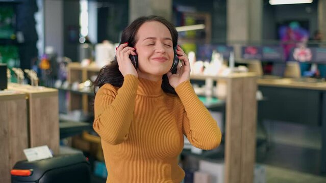 Attractive young girl with curly hair in an orange sweater is trying on headphones, posing for the camera and enjoying music