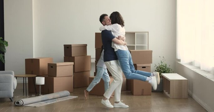 Happy family with daughter celebrate relocation, cheerful husband rise lifts spinning her wife joyful child running around feel overjoyed at move in day, heap of stuff nearby. Bank loan, life changes