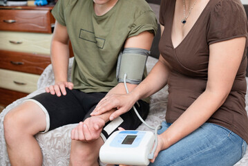 A woman measures her adult son's blood pressure with a torometer and measures pulse with hand, the...