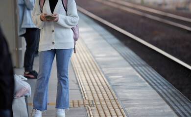 駅のホームで電車を待ちながらスマートフォンを弄る若い女性の姿 - obrazy, fototapety, plakaty