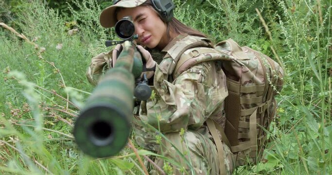 brave confident attractive caucasian woman soldier with sniper rifle with telescopic sight,keep defense in battle aims at enemy.camera motion military girl sitting in ambush,hiding in tall green grass