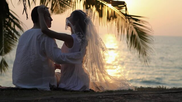 Newlyweds enjoy sunset against sun-path on ocean water. Bride in veil caresses groom under palm branch on beach after ceremony
