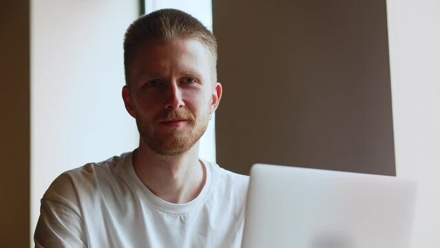 A frowning man working at a laptop turns to the camera and begins to smile. A man in a white t-shirt looks at something on the screen of his laptop.