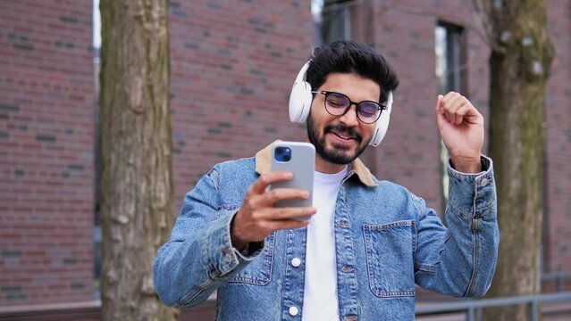 Smiling laughing handsome young indian man in glasses listening music from his smartphone in wireless white headphones, dancing on street, having rest free time enjoy good quality music outdoor.
