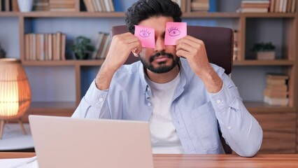 Sleeping Indian office worker is taking a humorous nap at his desk, using sticky notes to cover his eyes. The male employee appears tired and lazy, he seems to be pretending to work at office desk.