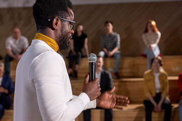 African lecturer presenting lesson with microphone to caucasian group of people. Young black teacher talking about finance in lecture hall. Side view on handsome guy in spectacles and white clothing
