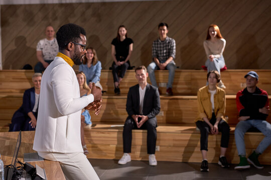 male african-american speaker giving presentation in lecture hall, audience. Young students in the background clapping hands. Conference event, training. Education, diversity, inclusive concept.