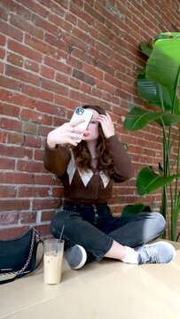 Lovely teen girl sitting on skateboard makes a selfies photo on the smartphone on a background red brick wall and large window. Hipster girl sits on a skateboard in front of red brick wall at modern