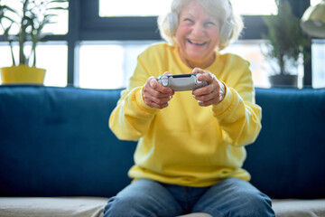 funny european senior woman with gray hair with joystick play online game at spare time in room interior, alone. Elderly people and modern gadget, retired people and modern technologies concept